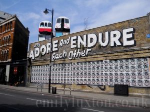 long brick wall with with two underground trains on top and large painted white words reading: "Let's Adore and Endure Each Other" - East London street art walk