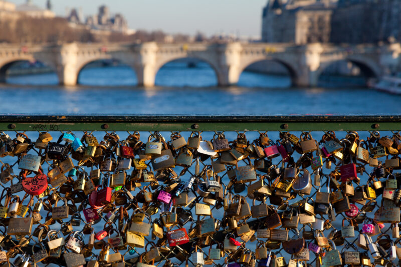 Hundreds of padlocks attached to a chain link fence on a bridge with a view of the river and a second stone bridge behind. 