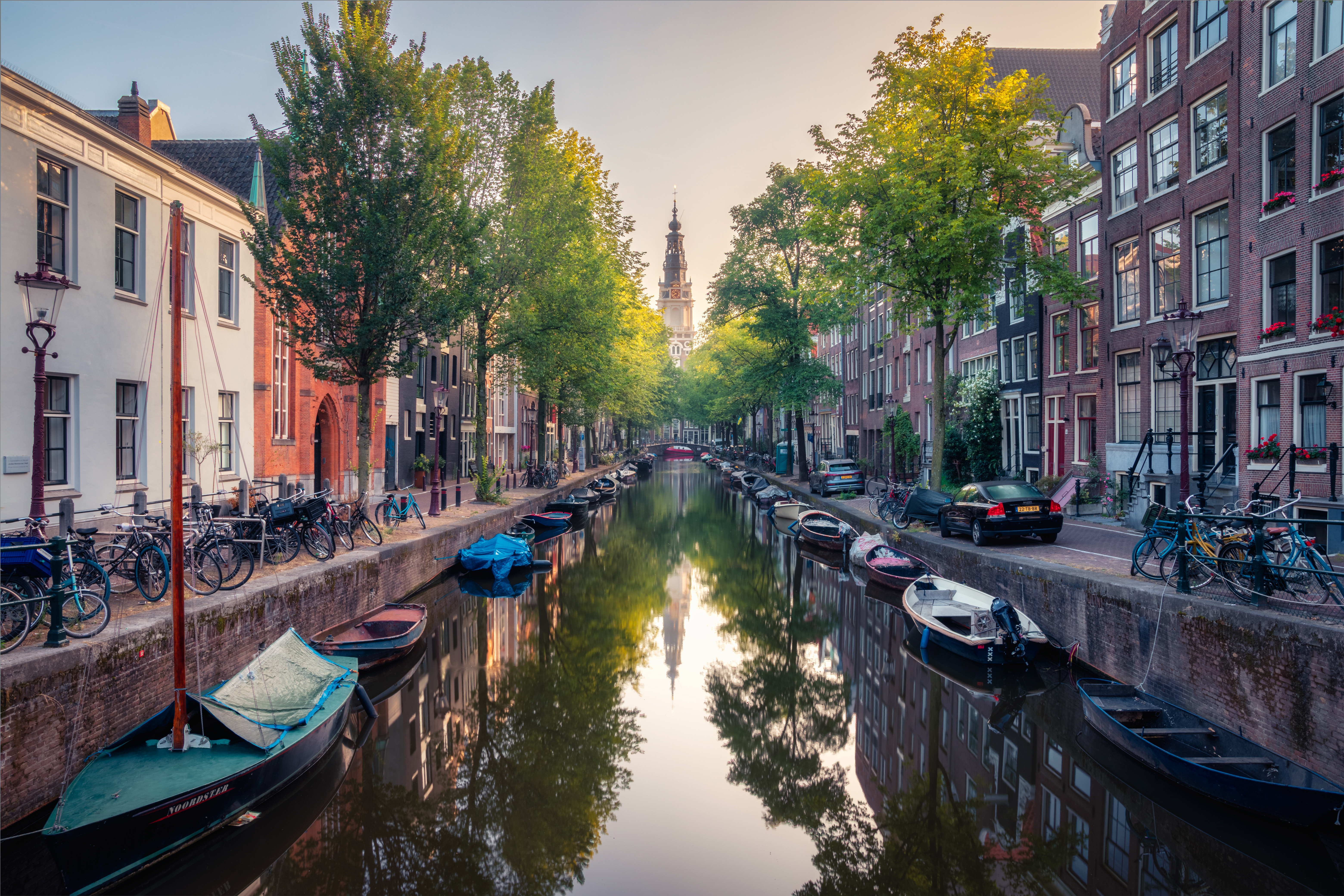 View along a canal towards the red brick, white topped tower of the Zuiderkerk church on a sunny day with leafy trees on either side and small boats along each edge of the canal. 