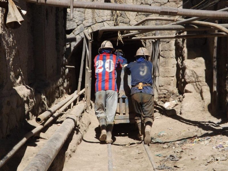 Two miners wearing dirty football shirrts and grey helmets, pushing a metal mine cart by hand along metal tracks towards a mine entrance with several pipes overhead and alongside. 