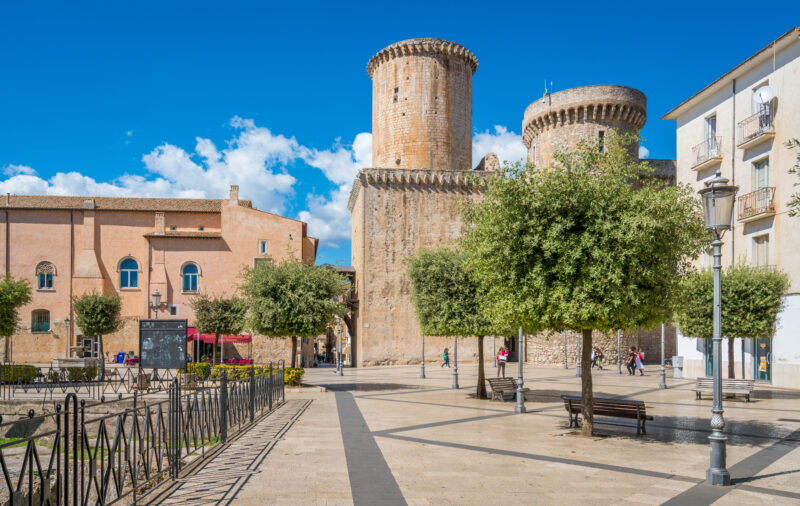 A picturesque town square featuring a historic castle with two large, cylindrical stone towers. The foreground has a paved plaza with trees, benches, and people walking around under a clear blue sky.