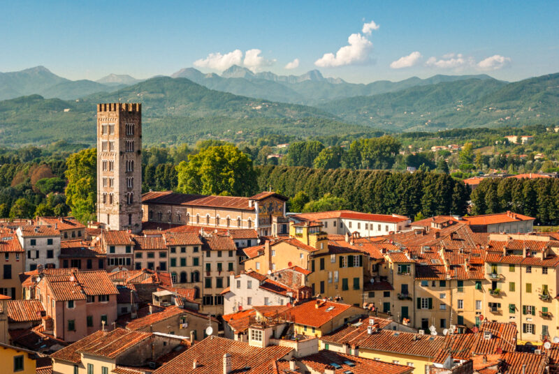 The Italian town of Lucca with medieval architecture, featuring a tall tower and red-tiled roofs, surrounded by lush green trees and rolling hills with distant mountains under a clear blue sky.