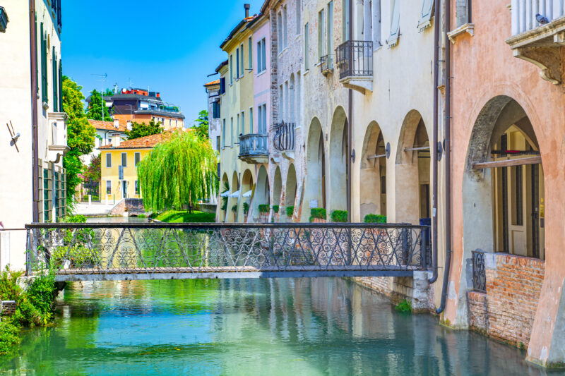 A picturesque canal in Treviso Italy flanked by pastel-colored buildings with arched walkways and balconies. A decorative iron bridge crosses the clear, reflective water. A lush green willow tree is in the background.