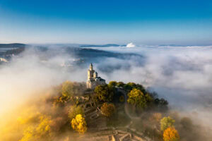 Aerial view of a hilltop church surrounded by dense fog and autumn trees, creating a mystical, ethereal atmosphere.