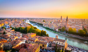 Aerial view of Verona cityscape at sunset, featuring a river with residential and historical buildings along its banks. Prominent tall towers and church spires indicate historical architecture.