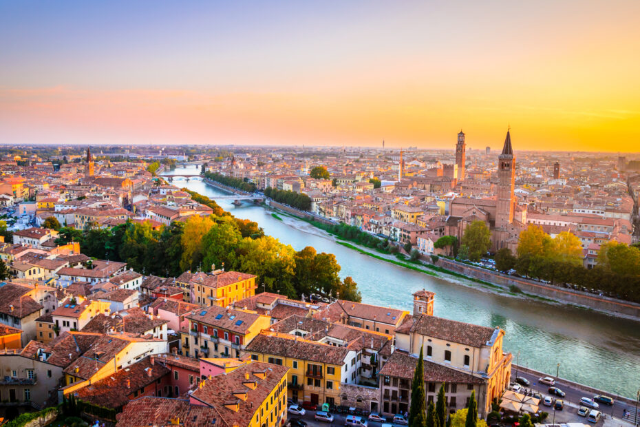 Aerial view of Verona cityscape at sunset, featuring a river with residential and historical buildings along its banks. Prominent tall towers and church spires indicate historical architecture.