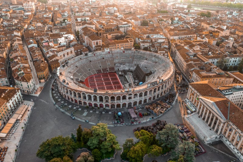 Aerial drone shot view of sunrise on ancient roman amphitheatre in Verona historic center, surrounded by urban streets and buildings - many with red tiled roofs.