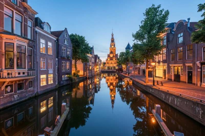 A picturesque canal scene in Alkmaar, with traditional Dutch buildings reflecting in the calm water. An illuminated clock tower stands in the background against a clear twilight sky, adding a focal point. Trees line the canal, enhancing the serene and charming atmosphere