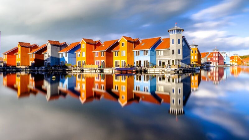 A row of colorful houses with red, blue, and gray exteriors along a calm waterfront, perfectly reflected in the still water. The sky is partly cloudy, adding to the serene atmosphere.