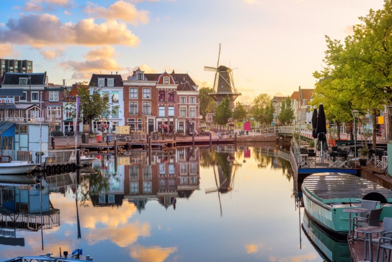 A picturesque in Leiden with traditional Dutch buildings and gabled roofs. A windmill stands in the background, and boats are docked along the calm water, reflecting the buildings and sky. The setting sun casts a warm, golden light.