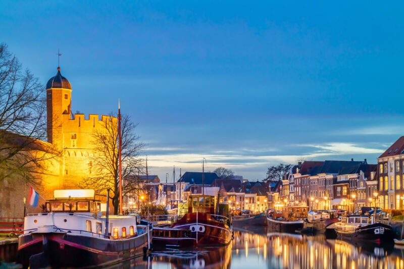 A canal in Zwolle at dusk lined with illuminated boats and historic buildings. A prominent brick tower with battlements and a conical roof stands on the left, adding a picturesque charm to the serene scene