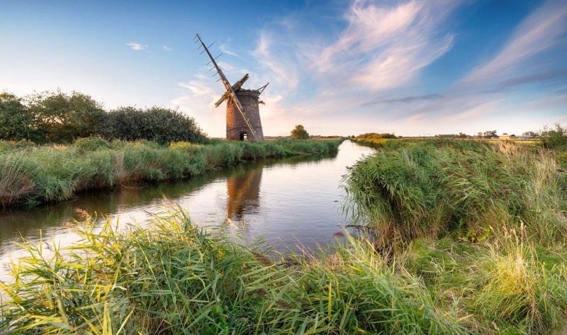 Windmill next to a calm flat river in the Norfolk Broads surrounded by grassland with a clear blue sky above