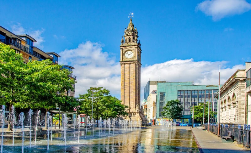 Albert Memorial Clock tower in Belfast, with a water fountain in the foreground and modern buildings in the background on a sunny day.