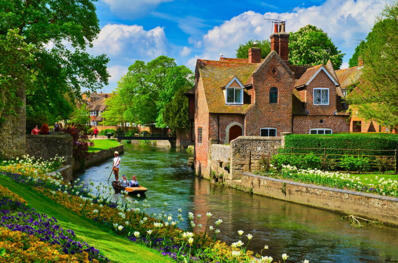 A small river flows through Canturbury with a brick house, colorful gardens, a stone bridge, and a person navigating a boat.