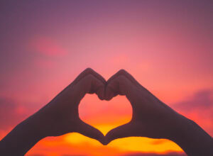 Silhouette of two hands creating a heart shape in front of a pink and orange sunset sky. Romantic Places in the UK.