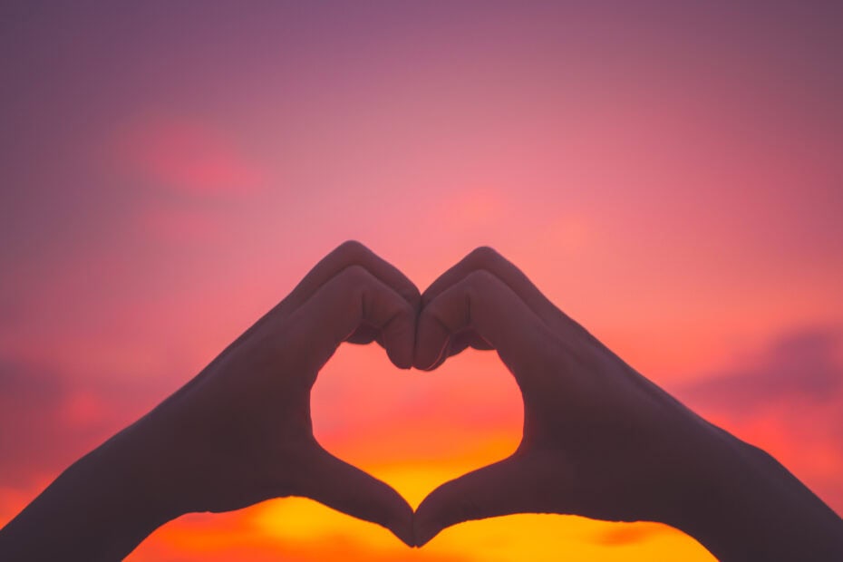 Silhouette of two hands creating a heart shape in front of a pink and orange sunset sky. Romantic Places in the UK.