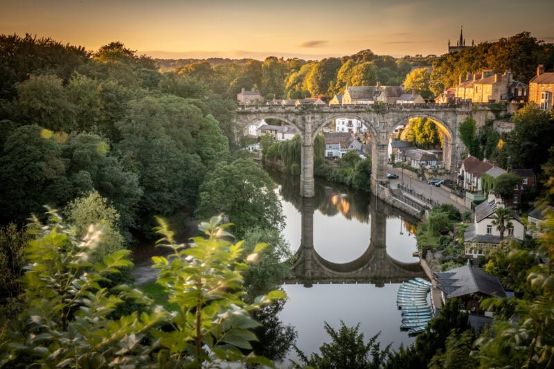 A stone viaduct spans over a calm river, reflecting its arches. The viaduct is surrounded by greenery and a small town with charming houses.