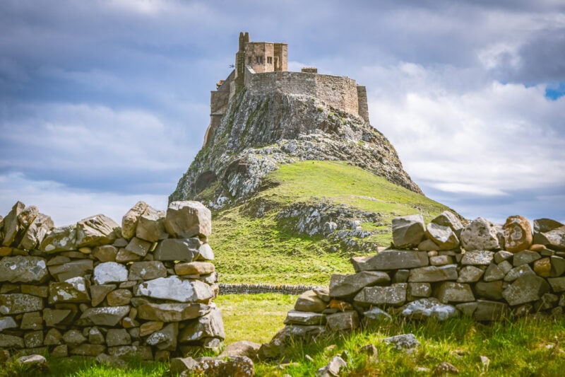 Lindisfarne Castle, a historic stone castle atop a steep, rocky hill, framed by a stone wall opening in the foreground, under a partly cloudy sky.