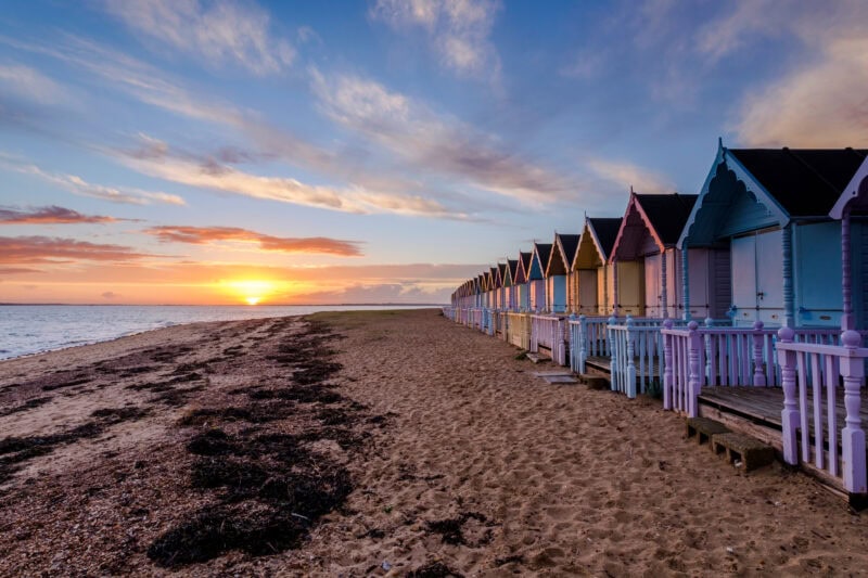 Colorful beach huts lined up along a sandy beach on Mersea Island at sunset, with a warm glow in the sky and seaweed on the sand.