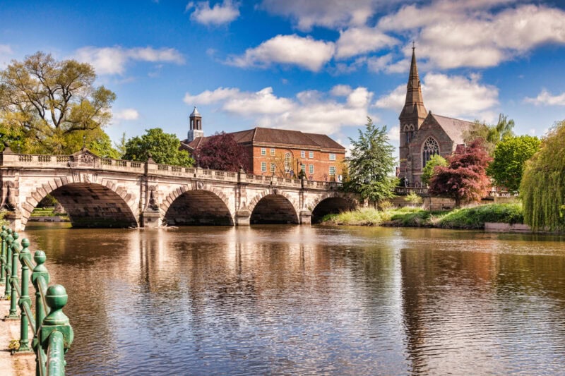 Arched stone bridge over a wide river with trees, a large red brick house and a church on the far bank.