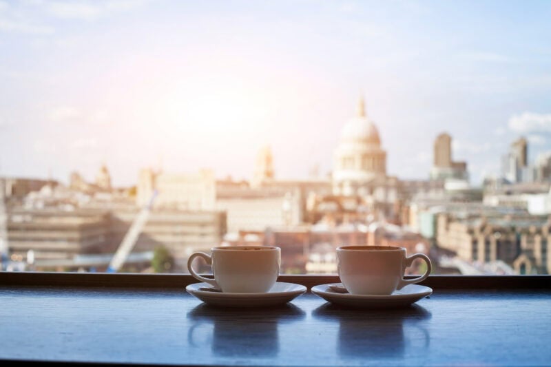 Two white cups of coffee on a table in front of a glass window with a view of St Paul cathedral and London skyline. Romantic Places in the UK. 