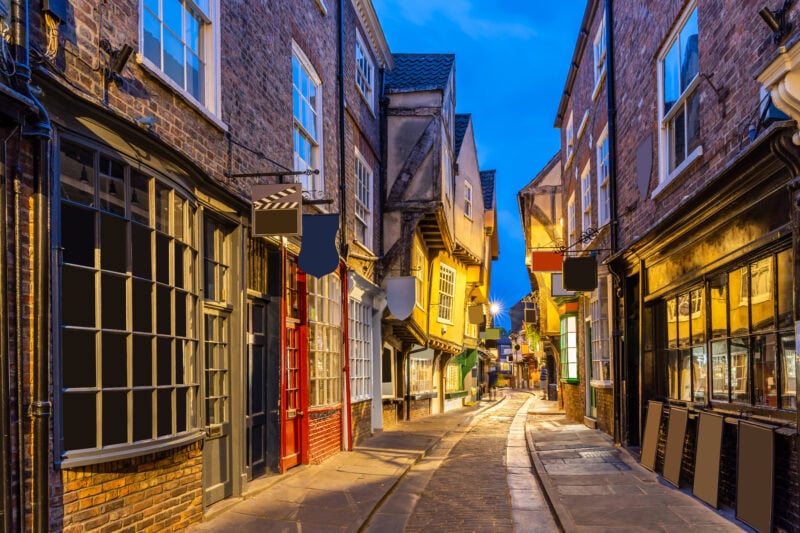 A narrow, cobblestone street with charming, historic buildings, large windows, and various shop signs, illuminated by warm, yellow lights.