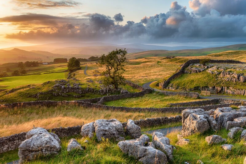 A picturesque countryside landscape at sunset with rolling hills, stone walls, a winding road, scattered trees, large rocks, and grassy fields.