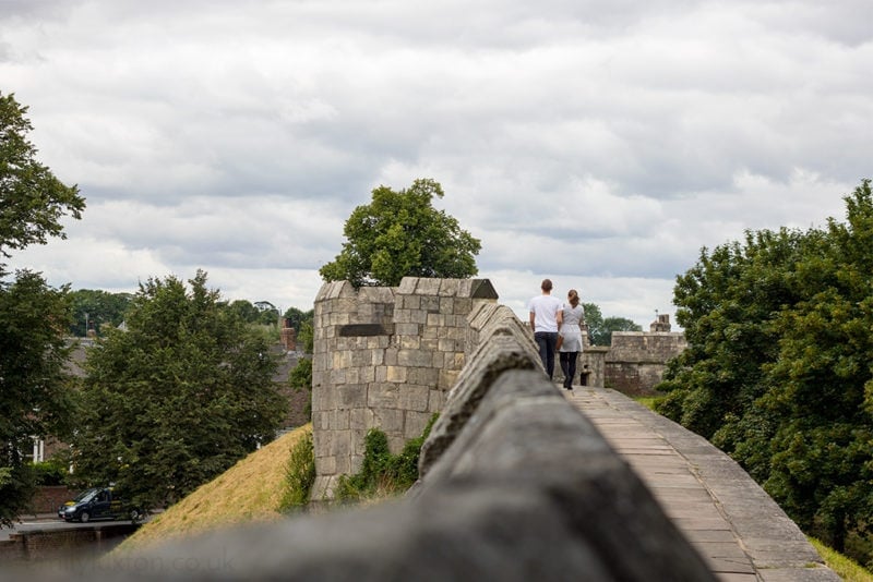 historic city wall built from grey stone with a low circular turret in the distance. a couple are walking along the wall holding hands in the distance, the man is wearing a white t shirt and the woman a denim jacket.