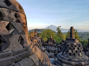 stone domes at Borobadur in Yogyakarta just after sunrise with a view of a conical mountain in the distance and blue sky overhead