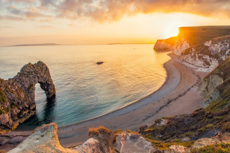 View from the edge of a grassy clifftop looking down at a bay with a curved shingle beach and a large rock archway over the sea. It is sunset and the sun is just going behind the cliffs with lens flare. Durdle door is one of the best things to do in dorset