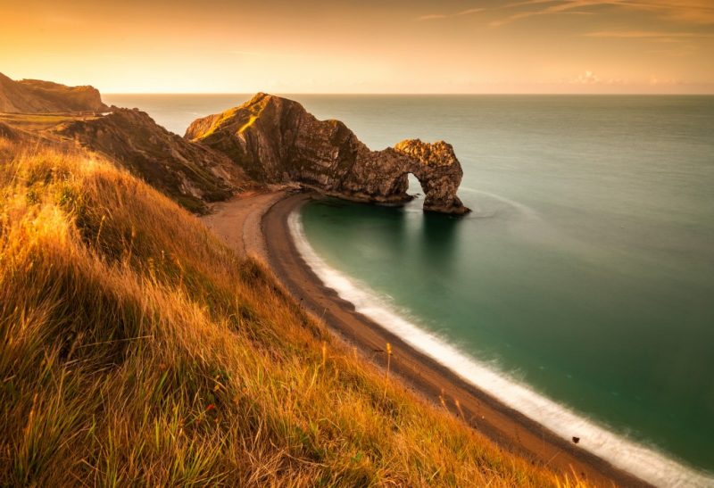 View of Durdle Door at sunset from a grassy clifftop looking down at the shingle beach and the large rock archway over the sea. Things to do in dorset