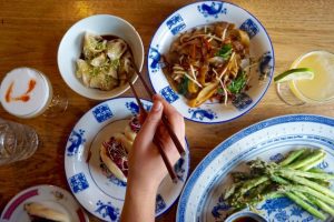 hand holding a set of chopsticks above several small bowls of food and a larger plate with grilled broccolli on a wooden table