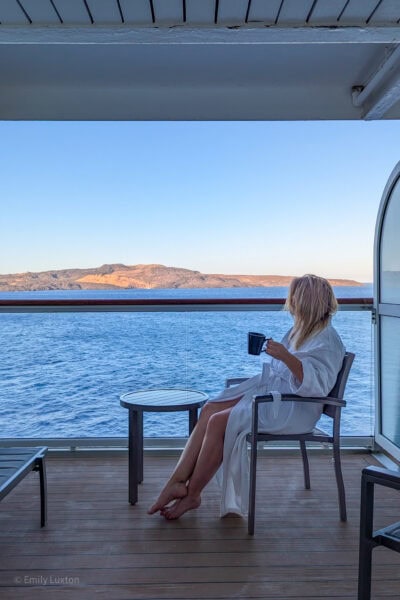 emily wearing a white dressing gown sitting alone on a grey garden chair next to a table on a cruise ship balcony with a view of the sea and a volcanic island near santorini just before sunset with golden haze and clear blue sky