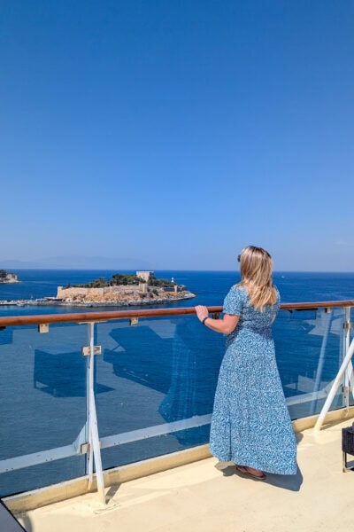 emily standing on the deck of a cruise ship looking away from the camera over the balcony towards blue sea and a small island with a castle on it. it is a very sunny day with clear blue sky above. 