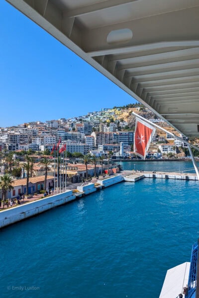 view of Kusadasi port from the rear of a cruise ship with a turkish flag hanging from the ship and bright blue water in the port. 