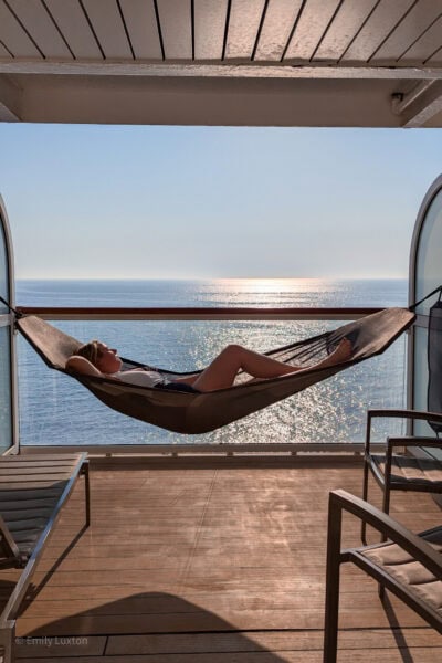 emily in a hammock on a balcony with a view of the blue sea behind. she is wearing a black and white swimsuit and has one leg raised up at the knee. 