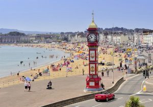 Red clock tower with a golden roof on a wide esplanade next to a sandy beach in Weymouth with a red sports car driving past