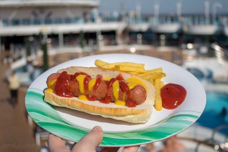 Close up of a hand hgolding a white and green plastic plate wiht a hot dog wiht zigzagged ketchup and mustard next to some french fries, held up in front of an outdoor cruise ship deck with the swimming pool out of focus in the background. From the Trident Grill on the Regal Princess cruise ship.