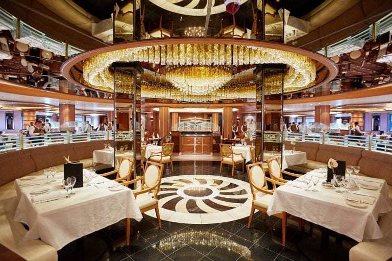 Interior of a large dining room on a cruise ship. there are several square tables with white tablecloths. the floor is black tiles with a white and black circle design in the middle underneath a huge gold and glass chandelier. the ciling is mirrored. Regal Princess Food Review