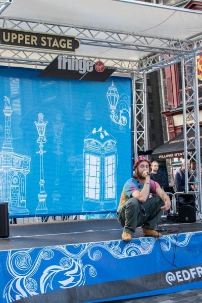 man in a tie-dye t-chirt and red bucket hat squatting on the edge of an outdoor festival stage singing into a microphone. Edinburgh fringe festival tips