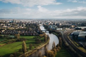 aerial view of York city with a river and green fields in th eforeground and a city skyline further back. There is a large cathedral taller than all the other buildings to the right and a cloudy sky above.