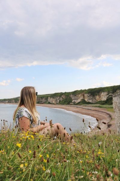 Emily wearing a white t shirt with palm trees printed on it, sitting down in the long grass and wildflowers at the top of the cliff and looking away from the camera towards a curved bay with a shingle and sand beach backed by low white cliffs