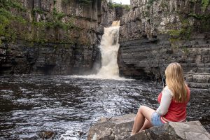 Emily wearing denim shorts and a red jumper with white sleeves, sitting on a rock by a river and looking away from the camera towards High Force Falls, a small and very powerfull waterfall with white water flowing down a dark grey rocky cliff face. Durham Dales