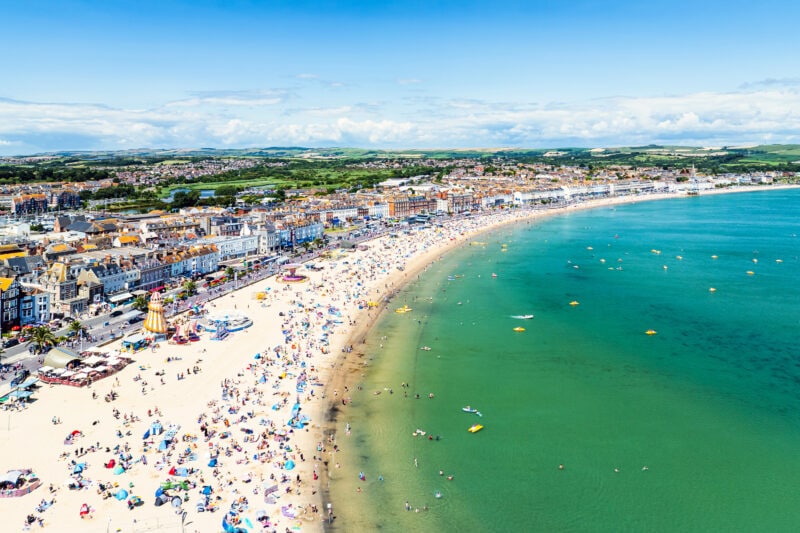 drone shot of weymouth bay, a white sandy beach crowded with people next to turquoise sea on a sunny summer day