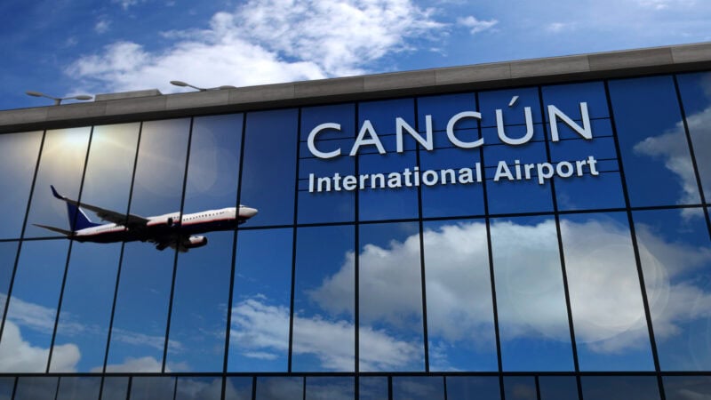 Reflection of an airplane descending through blue sky with fluffy clouds. This is reflected in a large glass building, with a sign in white letters reading: Cancun International Airport