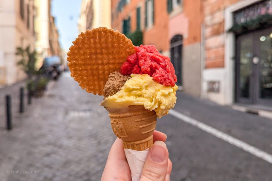 hand holding an ice cream cone with three different colour scoops of gleato: one red, one yellow and one brown. There is a circular wafer in the gelato and behind is a flagstone street in Rome lined with shops and orange buildings.