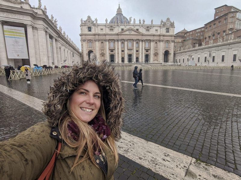 Emily taking a selfie in front of St Peter's Basilica in Vatican City on a rainy day with wet floor and grey sky. She is standing on wet grey cobblestones with the beige stone building of the basilica behind her and waring a khaki green coat with a large fur trimmed hood. 