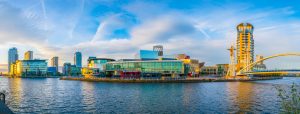 River with a city skyline on the far side with several waterfront buildings mostly made of glass. It is a very sunny day with blue sky above. places to visit in the north of england - manchester