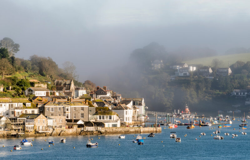 A picturesque coastal village with a collection of stone and brick houses situated on a hillside. Boats are moored in the calm water of the harbor. A misty hillside background adds a serene and atmospheric quality to the scene.