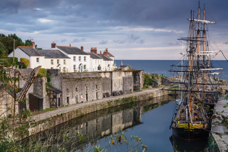 Historic coastal scene in Charlestown with a historic harbor, featuring white and beige houses with chimneys on a stone quay. A large classic sailing ship is docked in the calm harbor waters, reflecting the serene and historical atmosphere under a cloudy sky.